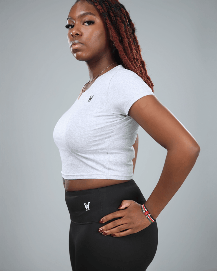 Woman wearing a light grey crop top and black leggings with logos on a gray background