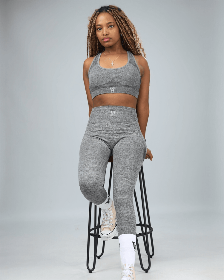Woman wearing a grey sports bra with waaka logo on a plain background sitting on stool
