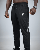 Black jogger pants with a logo on the leg worn by a person against a gray background