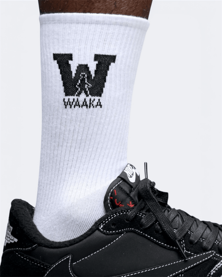 White sock with black waaka logo and text worn with a black sneaker on a light gray background