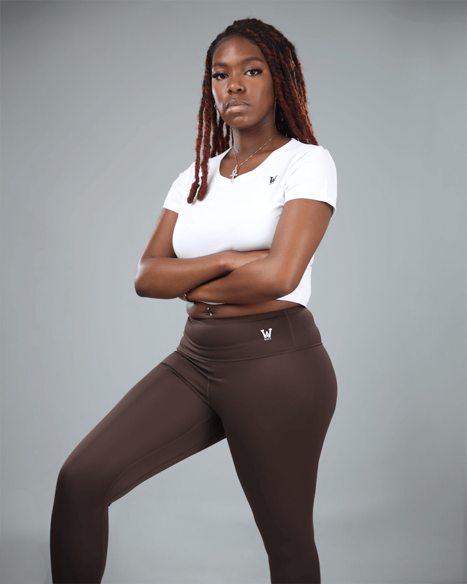 Woman wearing a white t-shirt and brown leggings with waaka logo on a gray background with arms folded