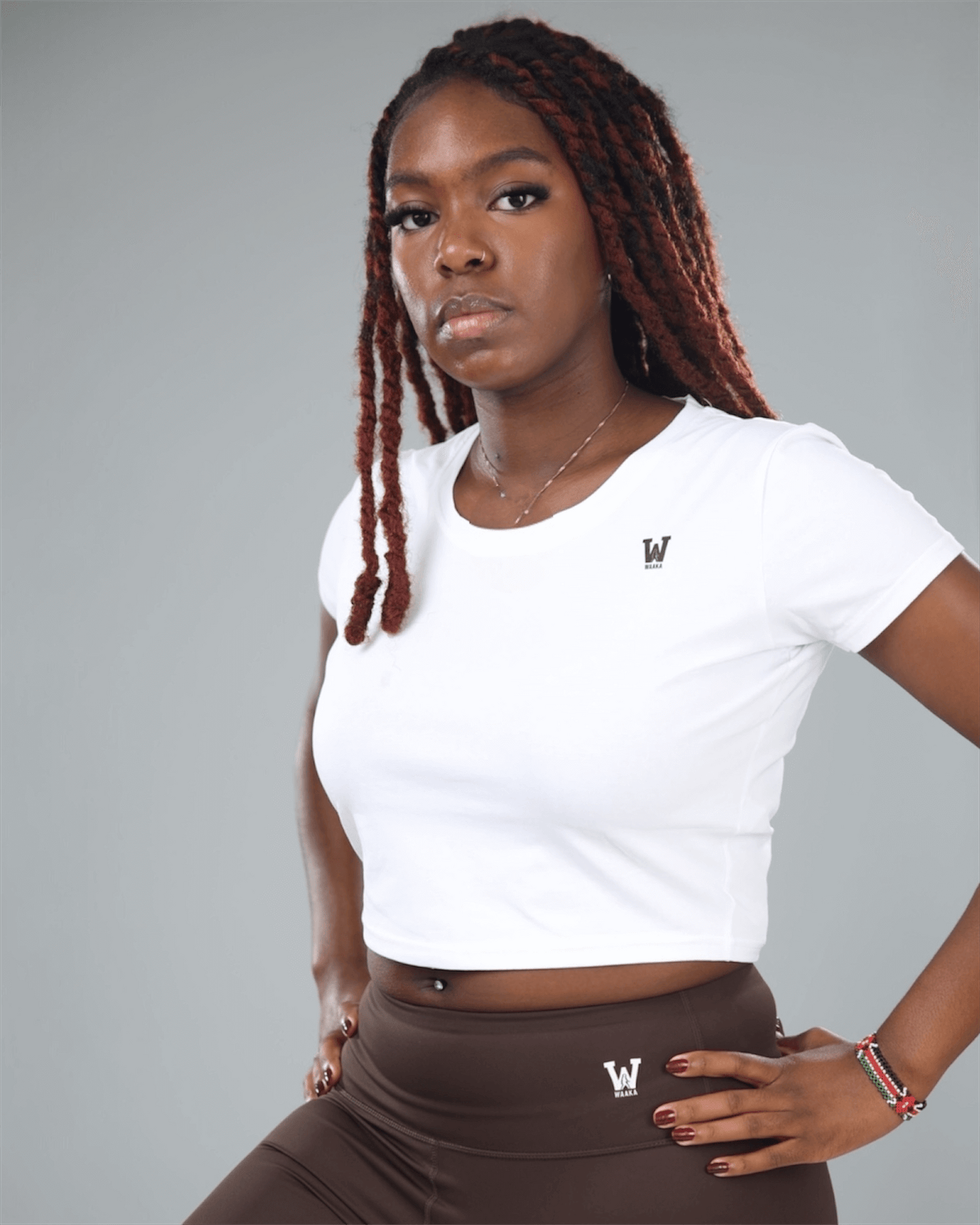 Woman wearing a white crop top and brown skirt with waaka logo, standing against a gray background
