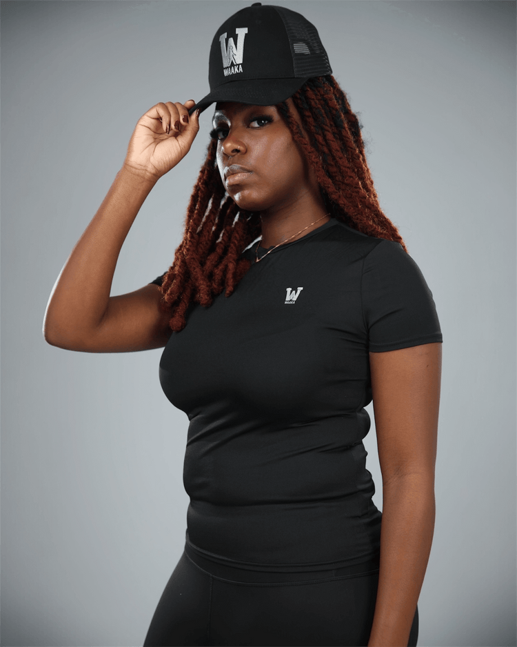 Woman wearing a black short sleeve  t-shirt with waaka logo on a grey background with a black cap