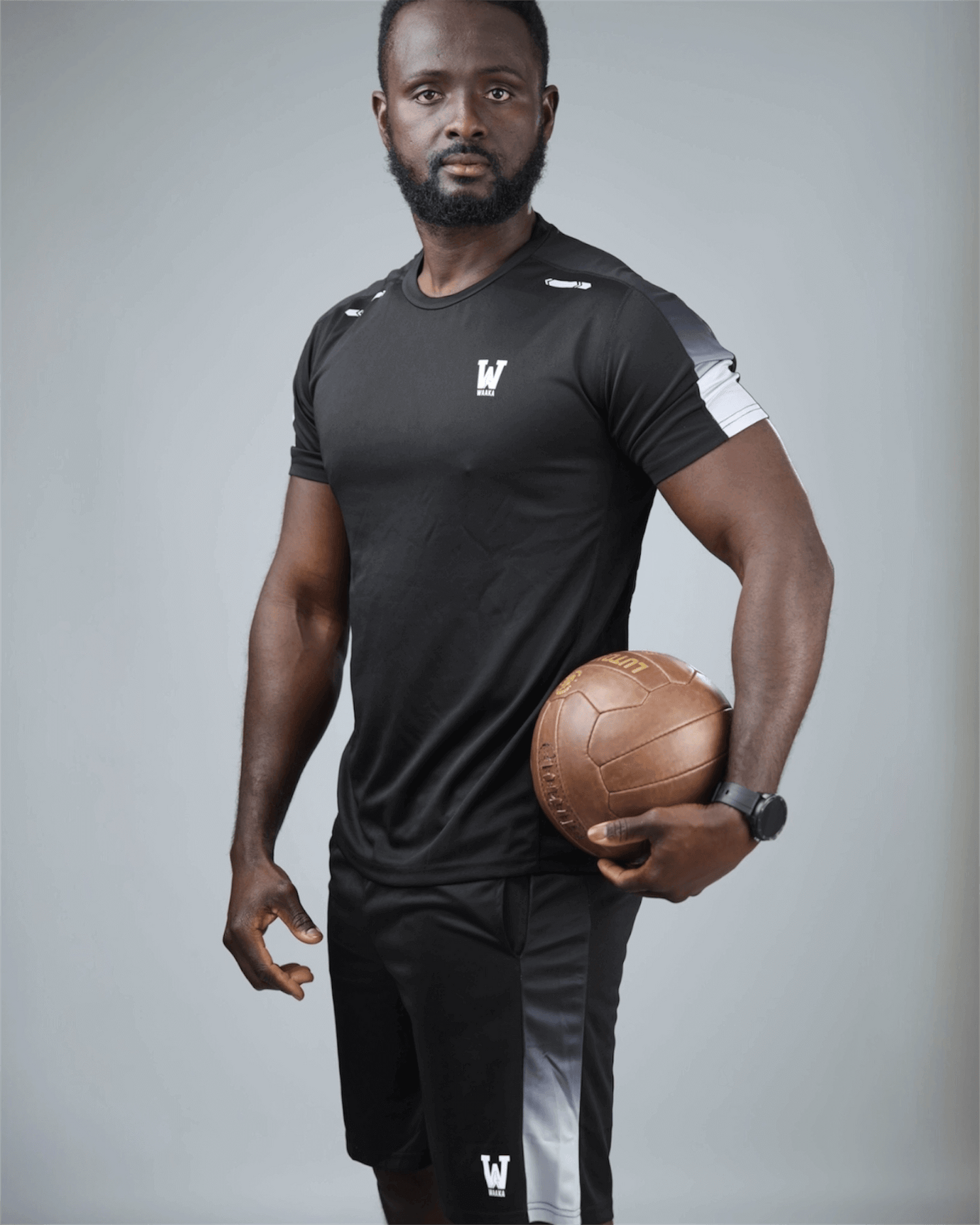 Man wearing a black athletic outfit with a logo, holding a medicine ball against a gray background