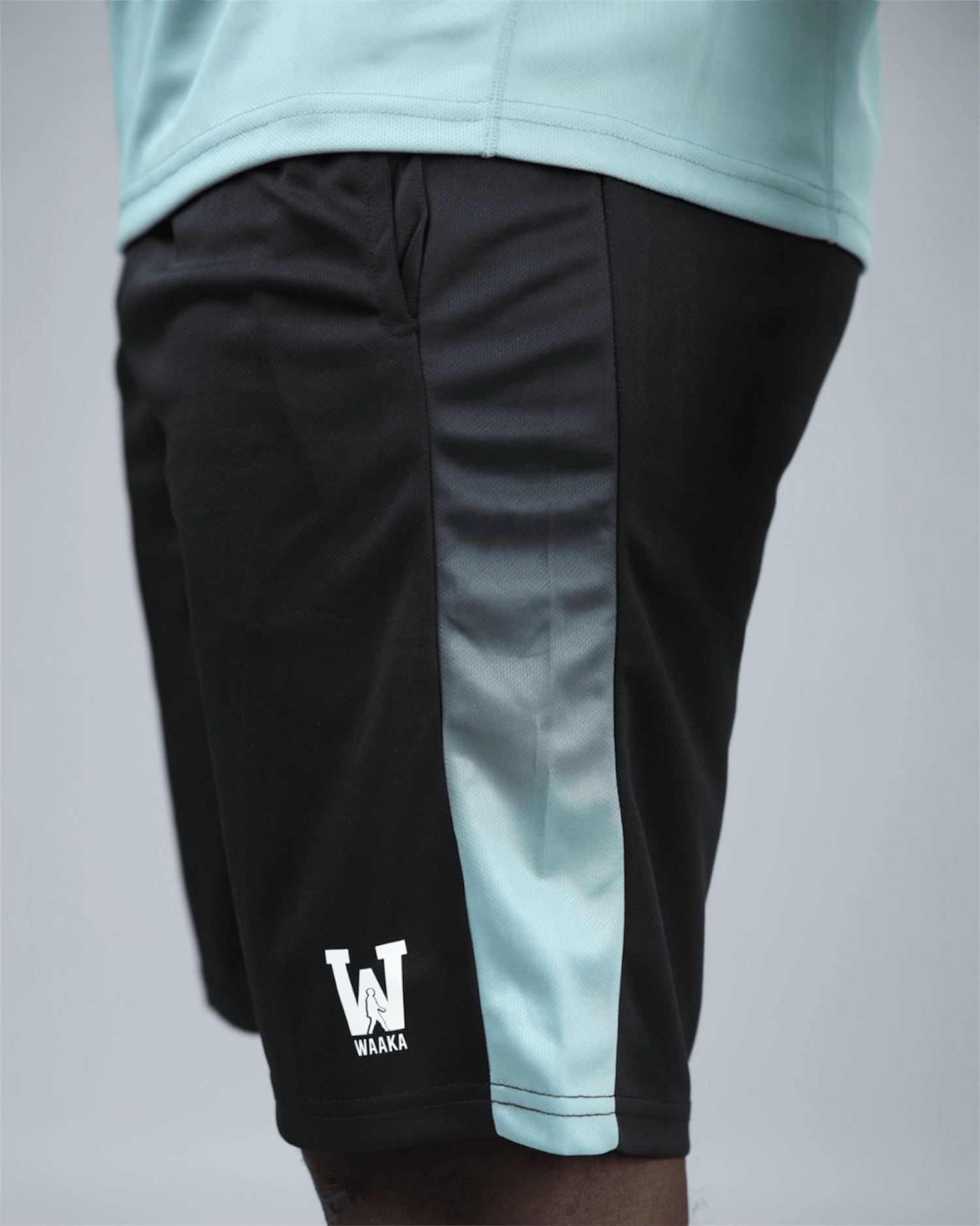 Black eco training shorts with a light blue stripe featuring waaka logo on a grey background