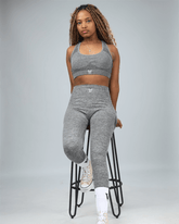 Woman wearing a grey sports bra with waaka logo on a plain background sitting on stool